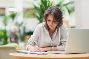 An independent student writing on a pad in front of a laptop